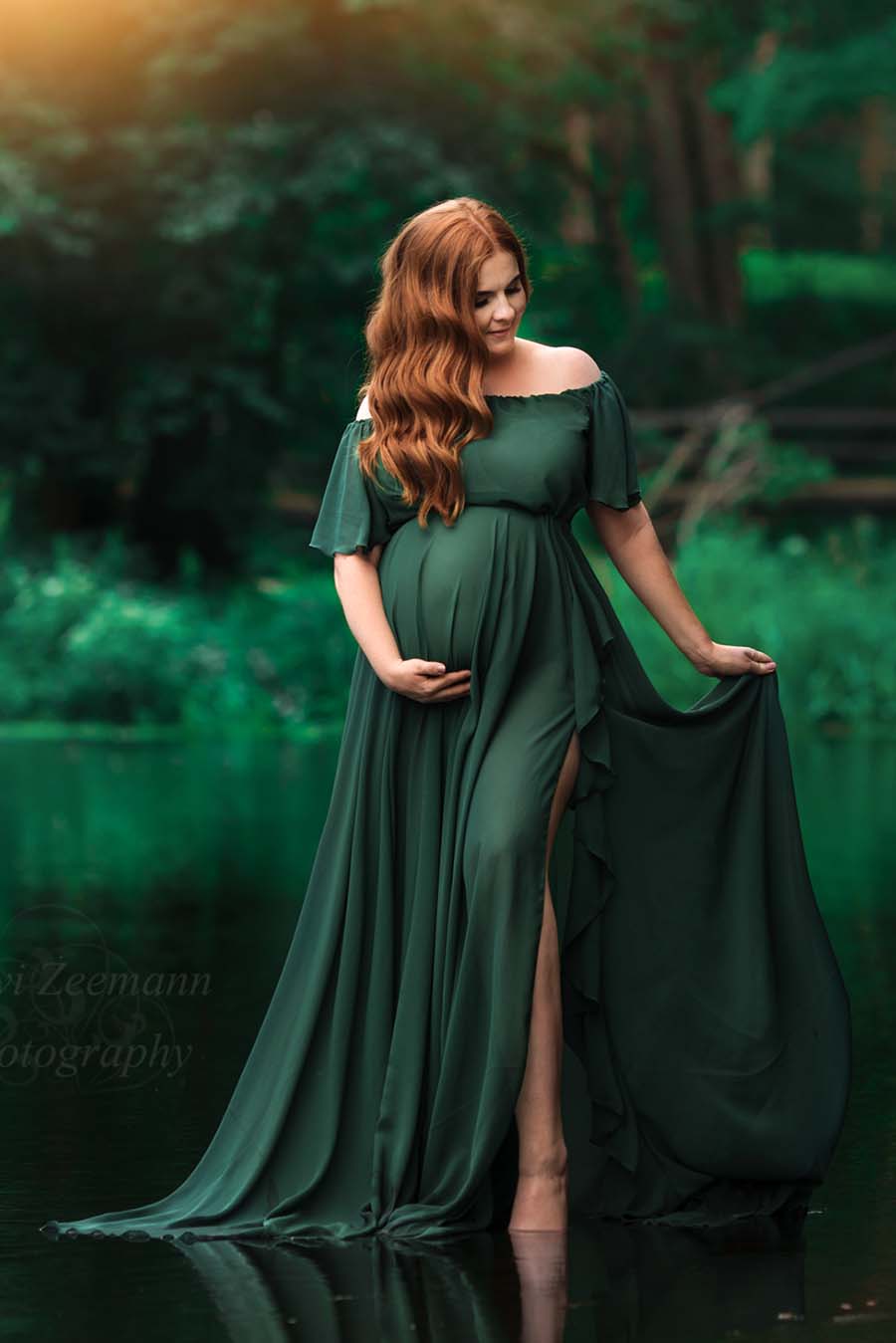 red haired pregnant model poses by the water wearing a chiffon dark green dress with a long circle skirt - the skirt has a delicate split on the side with ruffles at the ending. the top is off shoulder and has short ruffle sleeves. 