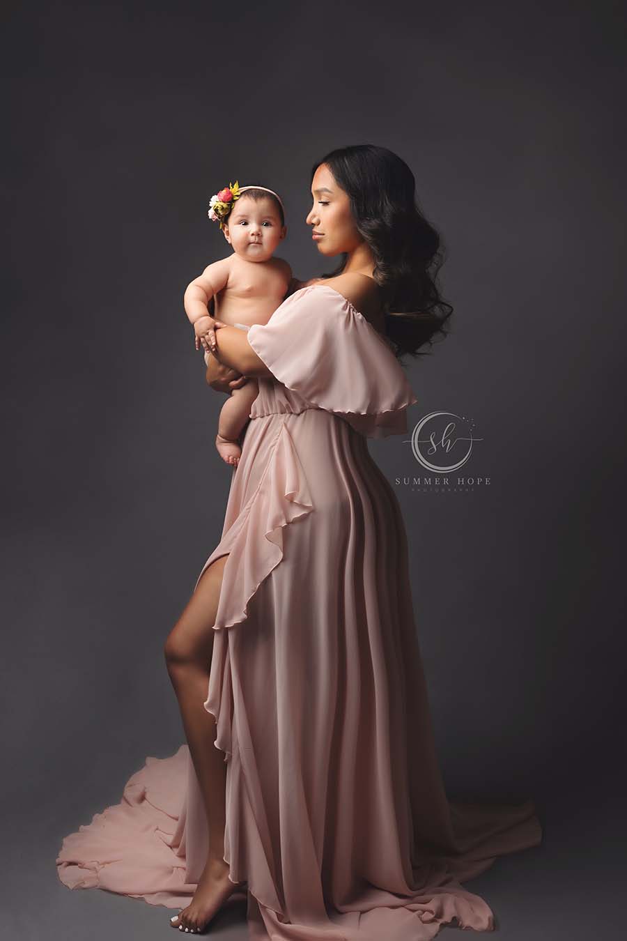 brunette model poses in a studio holding her baby girl and wearing a dusty pink chiffon long dress with a ruffle top and a split on the side. 