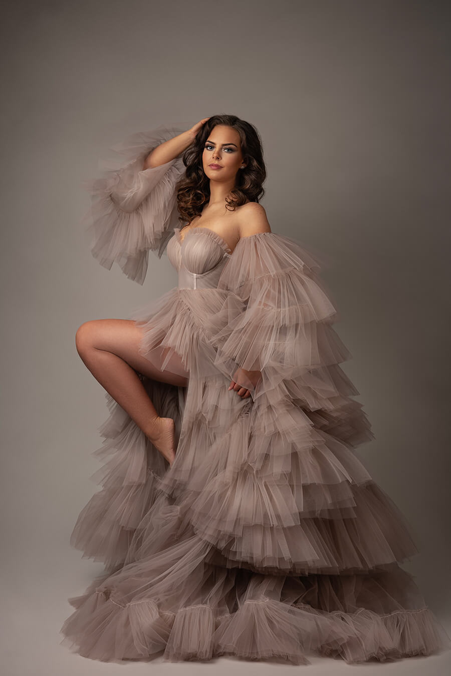 brunette model poses in a studio wearing a large tulle dress in sand color. there is a split on the skirt, and a lot of tulle in the sleeves.