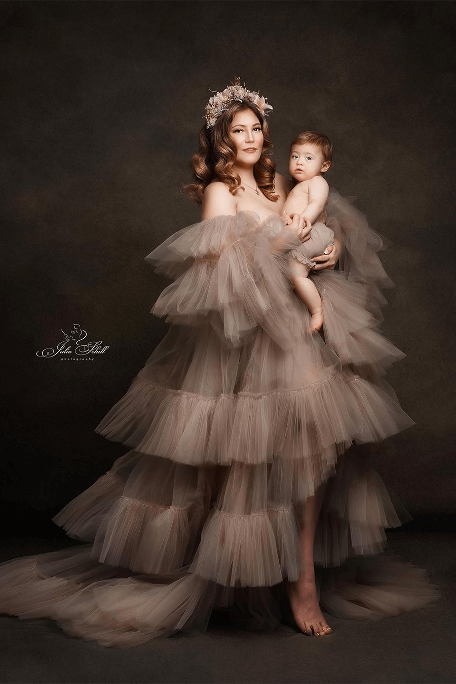 mother poses in a studio together with her kid. she is wearing a large tulle dress in sand color and a flower crown on her head.
