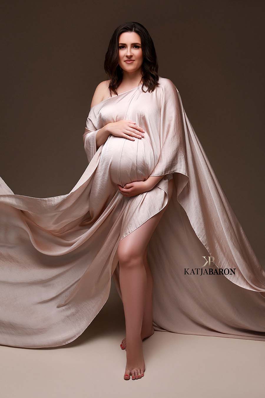 A pregnant model is wearing a cape. The cape has a hole in the middel where you can put your head trough. The fabric is hanging around her and has little waves in it. She has her hands around her belly. 