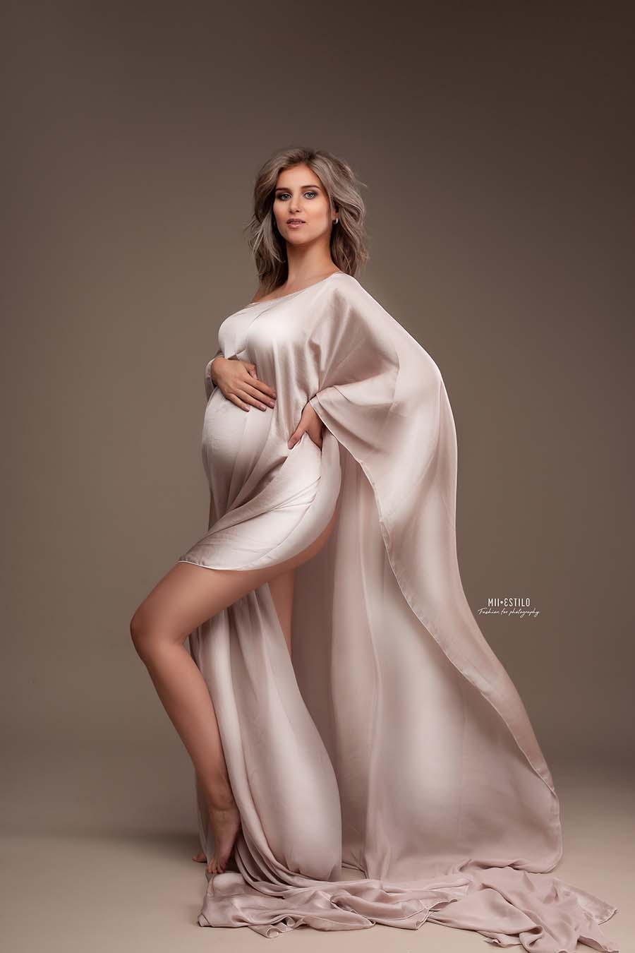 A pregnant model is posing in our amina cape sand. The material of the cape is silky for that nice shiny look. The model has short blonde hair. The fabric is hanging around her and it looks a little big like she is wearing a dress. 