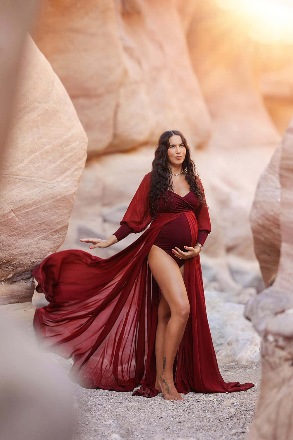 Pregnant model with long curled hair poses outside wearing a maternity outfit made of jersey and chiffon. The dress color is bordeaux and it features an adjustable sweetheart neckline and long chiffon bishop sleeves with jersey manchettes. the chiffon skirt has a split on the middle and a jersey bodysuit can be seen underneath. 