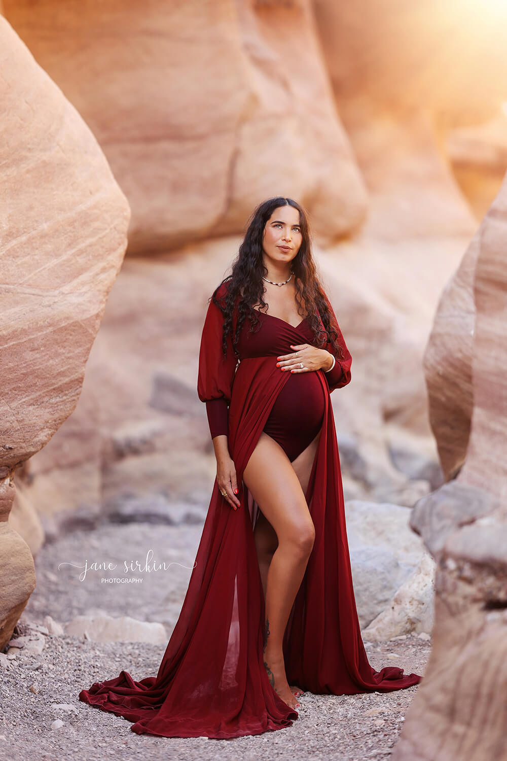 pregnant model with long curled hair poses outside wearing a maternity outfit made of jersey and chiffon. The dress color is bordeaux and it features an adjustable sweetheart neckline and long chiffon bishop sleeves with jersey manchettes. the chiffon skirt has a split on the middle and a jersey bodysuit can be seen underneath.