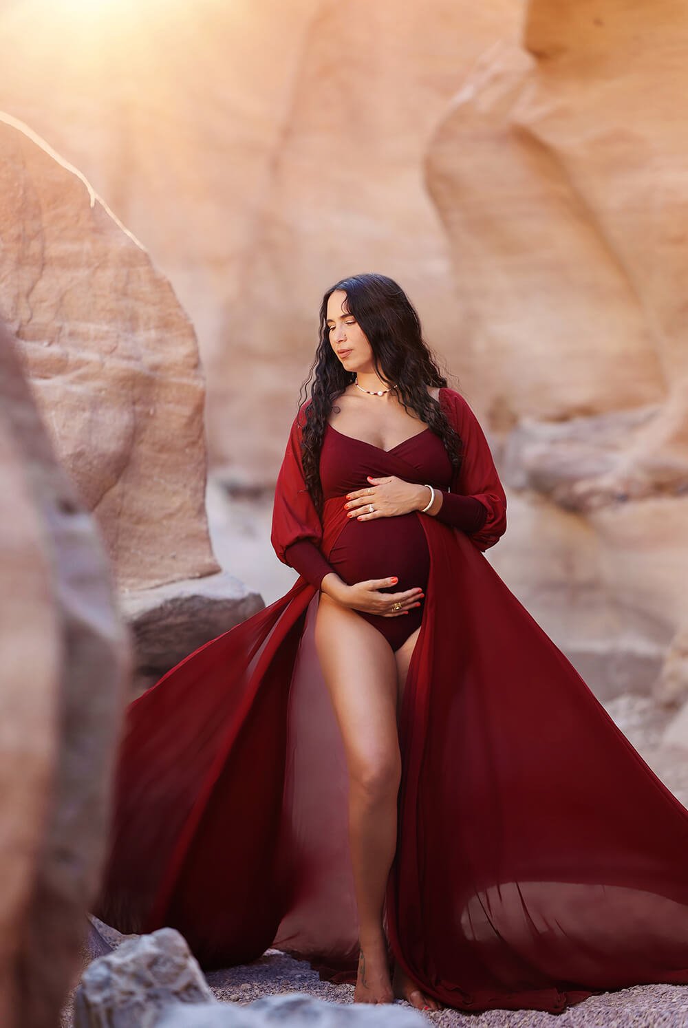 Pregnant model with long curled hair poses outside wearing a maternity outfit made of jersey and chiffon. The dress color is bordeaux and it features an adjustable sweetheart neckline and long chiffon bishop sleeves with jersey manchettes. the chiffon skirt has a split on the middle and a jersey bodysuit can be seen underneath.