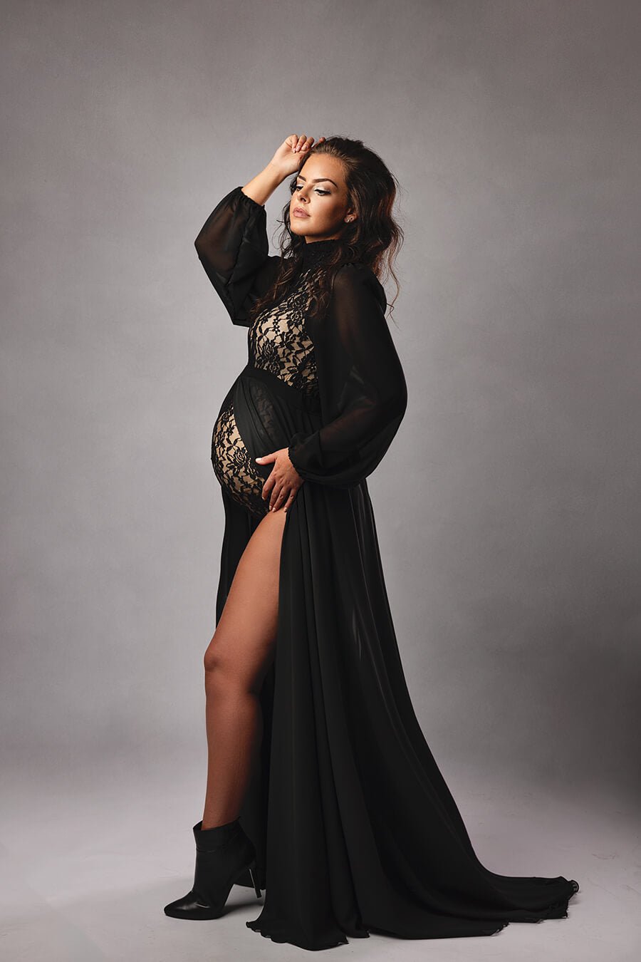 brunette pregnant model poses in a studio wearing a black lace bodysuit with chiffon sleeves and a long chiffon skirt with a split