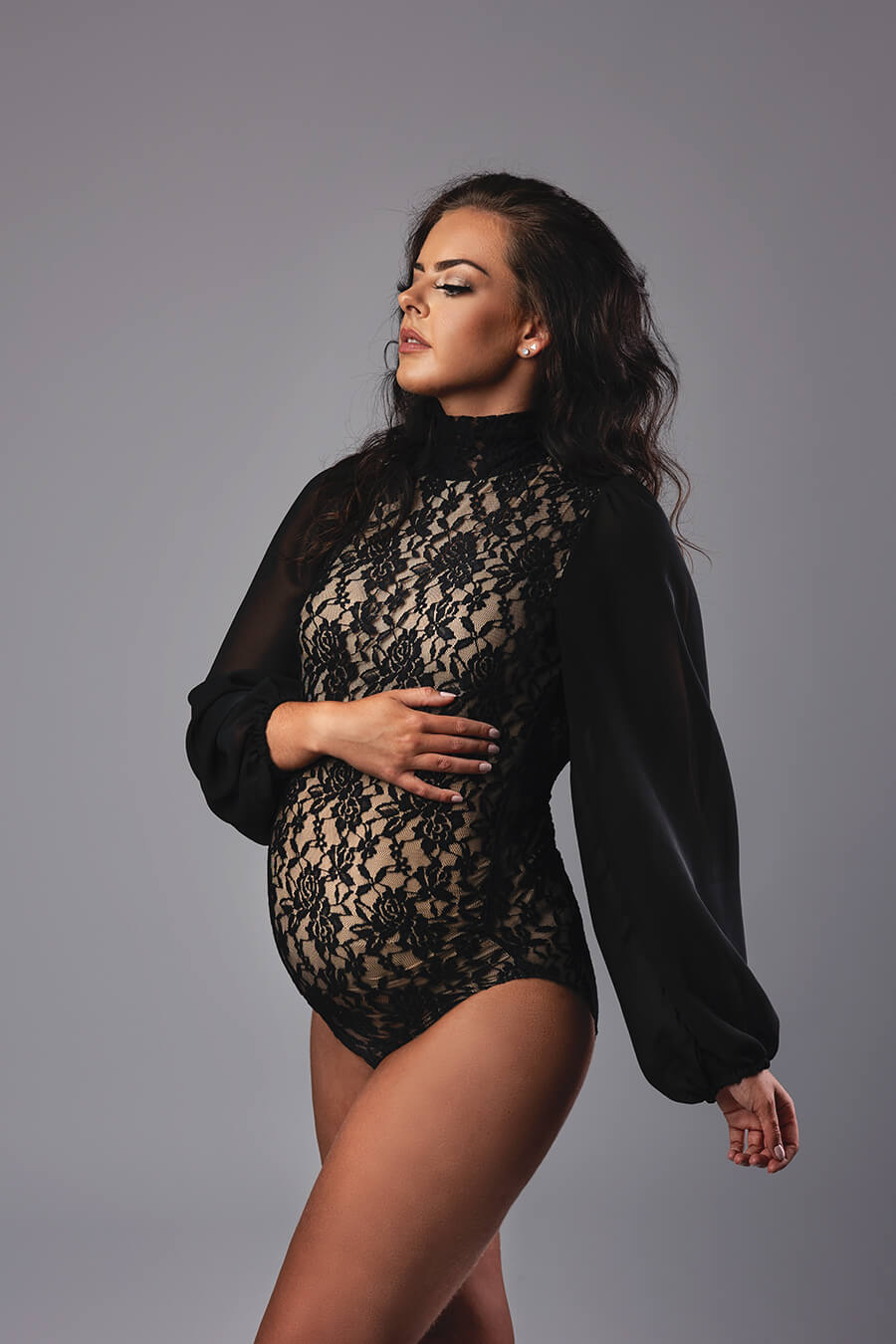 brunette pregnant model poses in a studio wearing a black lace bodysuit with chiffon bishop sleeves 