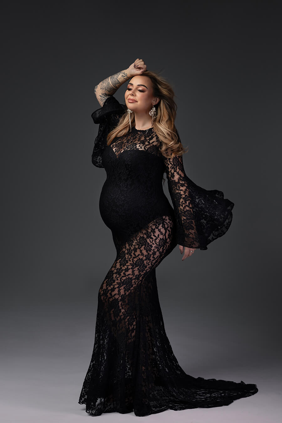 blond pregnant model poses in a studio wearing a long lace dress with bell sleeves and a bodysuit under