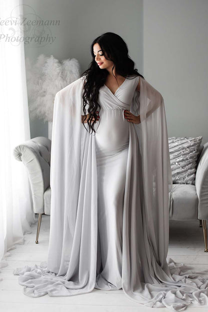 A model is standing in a living room setting. The room has a soft grey color. The model has long black hair and is wearing a long dress. The dress has loose chiffons material. by the shoulders that ends at the ground. It has a V neckline.