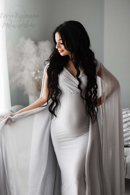 A pregnant woman is wearing a coolgrey dress. The dress is a tight fit model with loose chiffon grey fabric. 