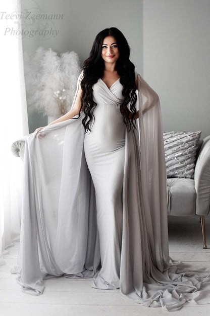 The model is a pregnant woman with long black har. She is smiling towards the camera. In the background there is a white plant and a grey couch. The dress that she is wearing is long and in a coolgrey color. She is holding a piece of the chiffon fabric. The dress is made out of jersey
