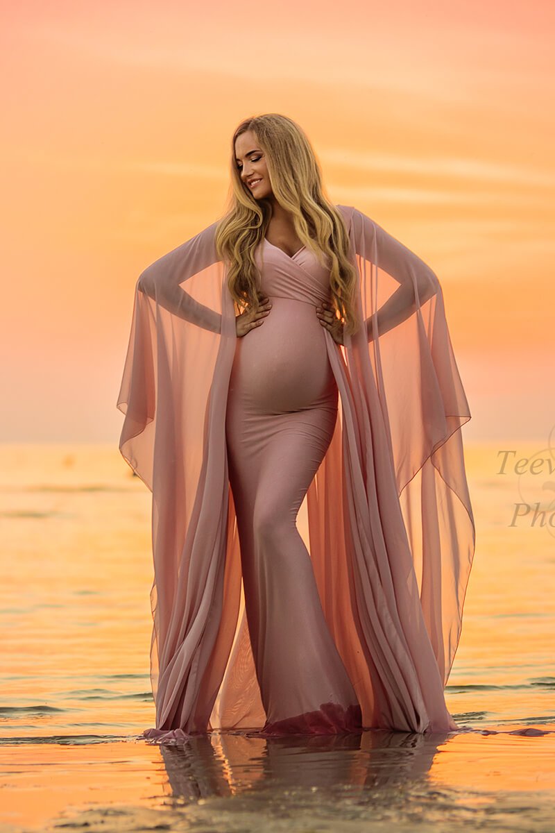 The model is standing in a beautiful long pink dress with loose sleeves. It is sunset