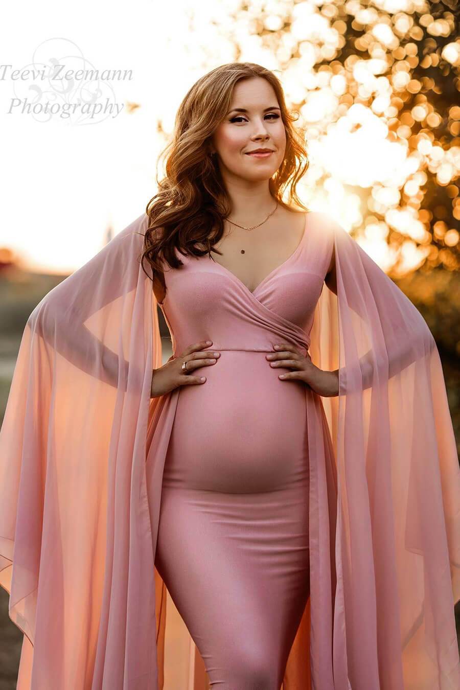 This pregnant women has short brown curly hair. She is wearing a long tight pink dress. The sleeves are loose and made out of chiffon
