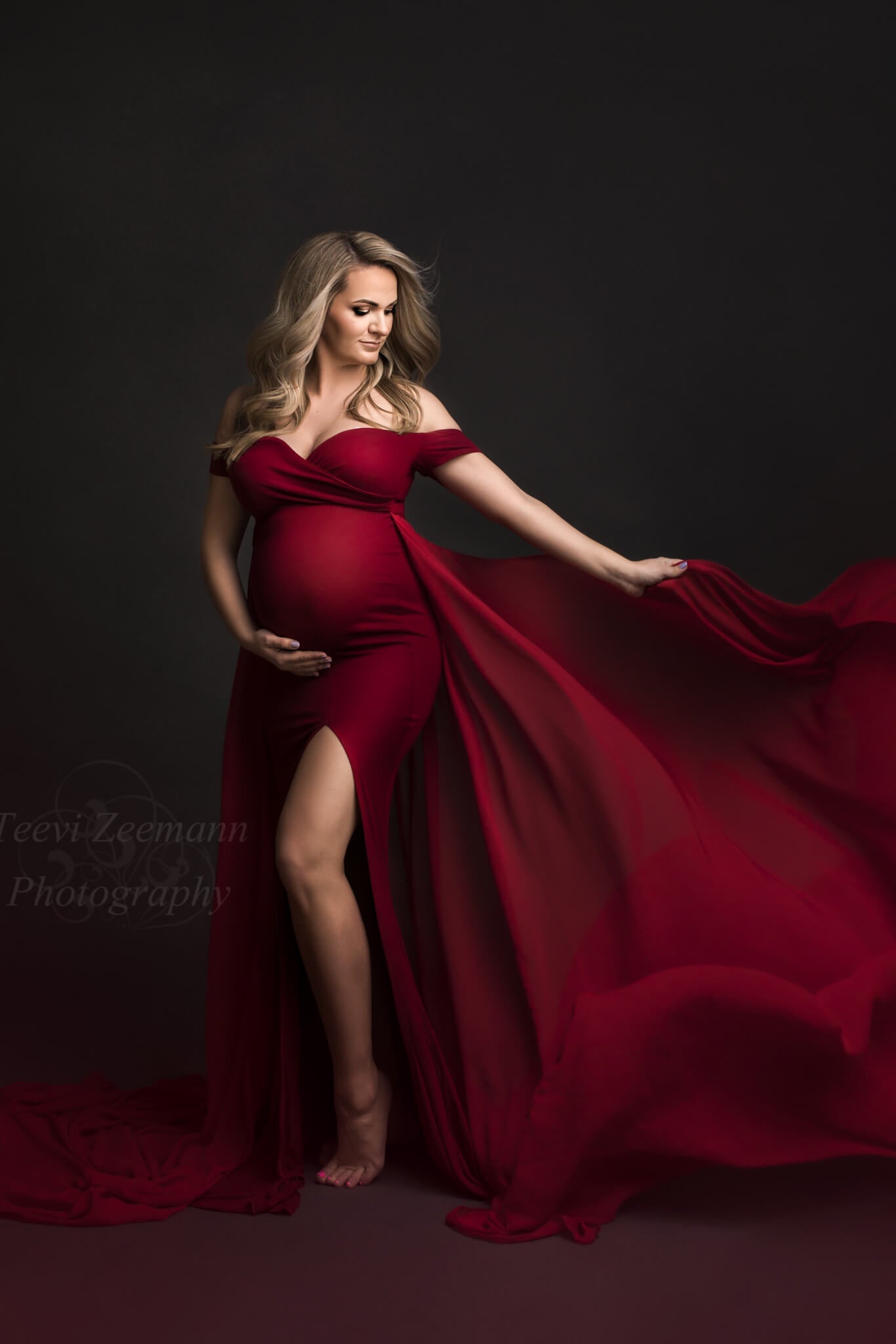 Blond pregnant model poses in a studio during a maternity photoshoot. She holds her bump with one hand while playing with the train from the dress with the other one. She wears a red dress with a side split and chiffon train.