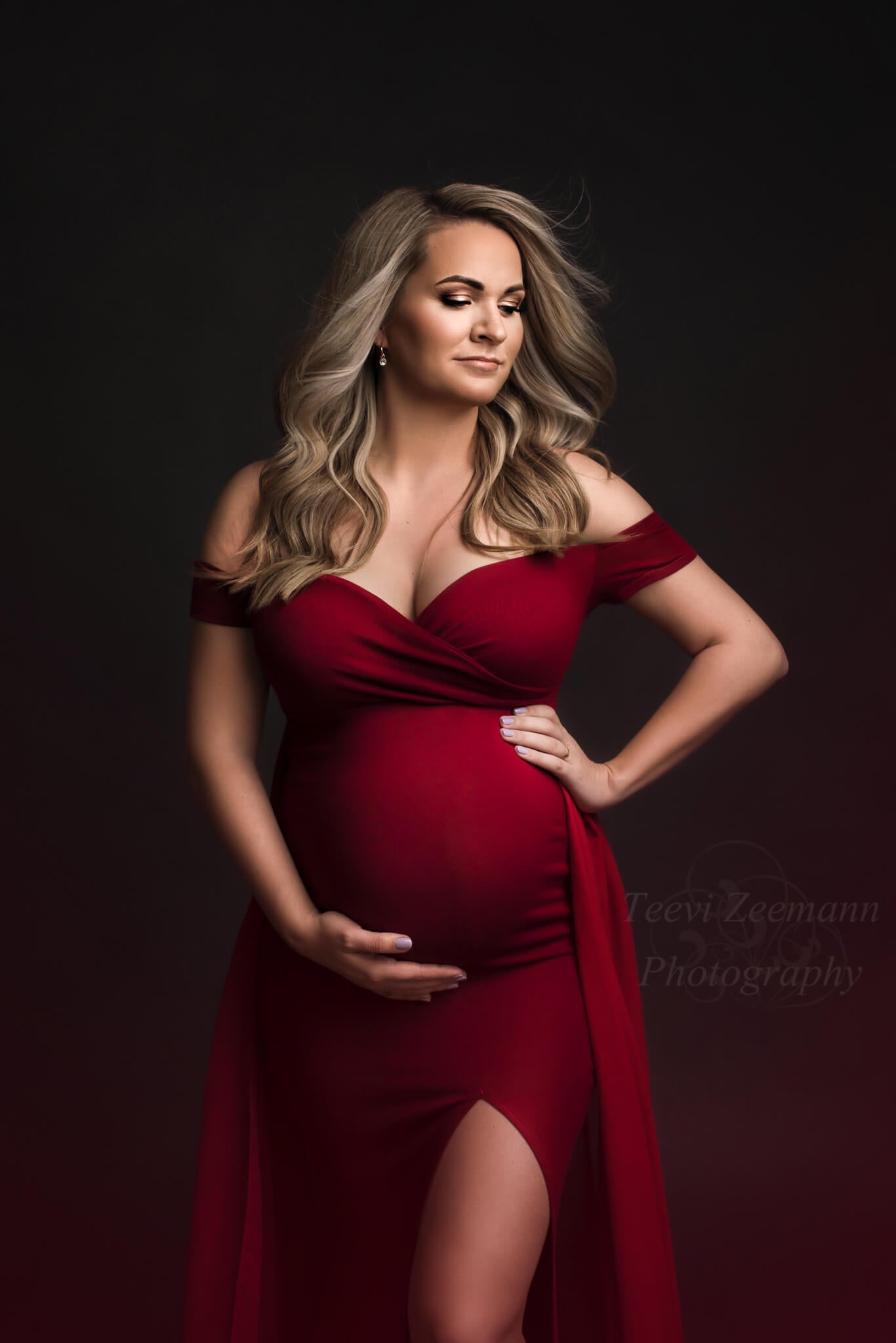 Blond pregnant model poses in a studio during a maternity photoshoot. She holds her bump with both hands. She wears a red dress with a sweetheart neckline and short sleeves. In the close up she has her eyes partially closed and stares to the side. 