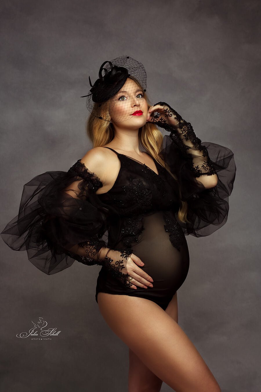 blond pregnant model poses in a studio wearing a special black bodysuit with 3d bridal lace. the bodysuit features tulle bishop sleeves and gloves. the model has a black piece on her head. 