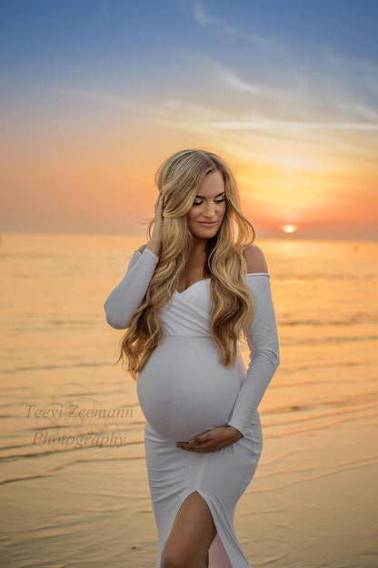 Blond pregnant model poses at the beach wearing a long tight white dress with a split on the skirt, long sleeves and sweetheart top. Model looks away and poses on her side. She holds her belly with one hand and her head with the other.