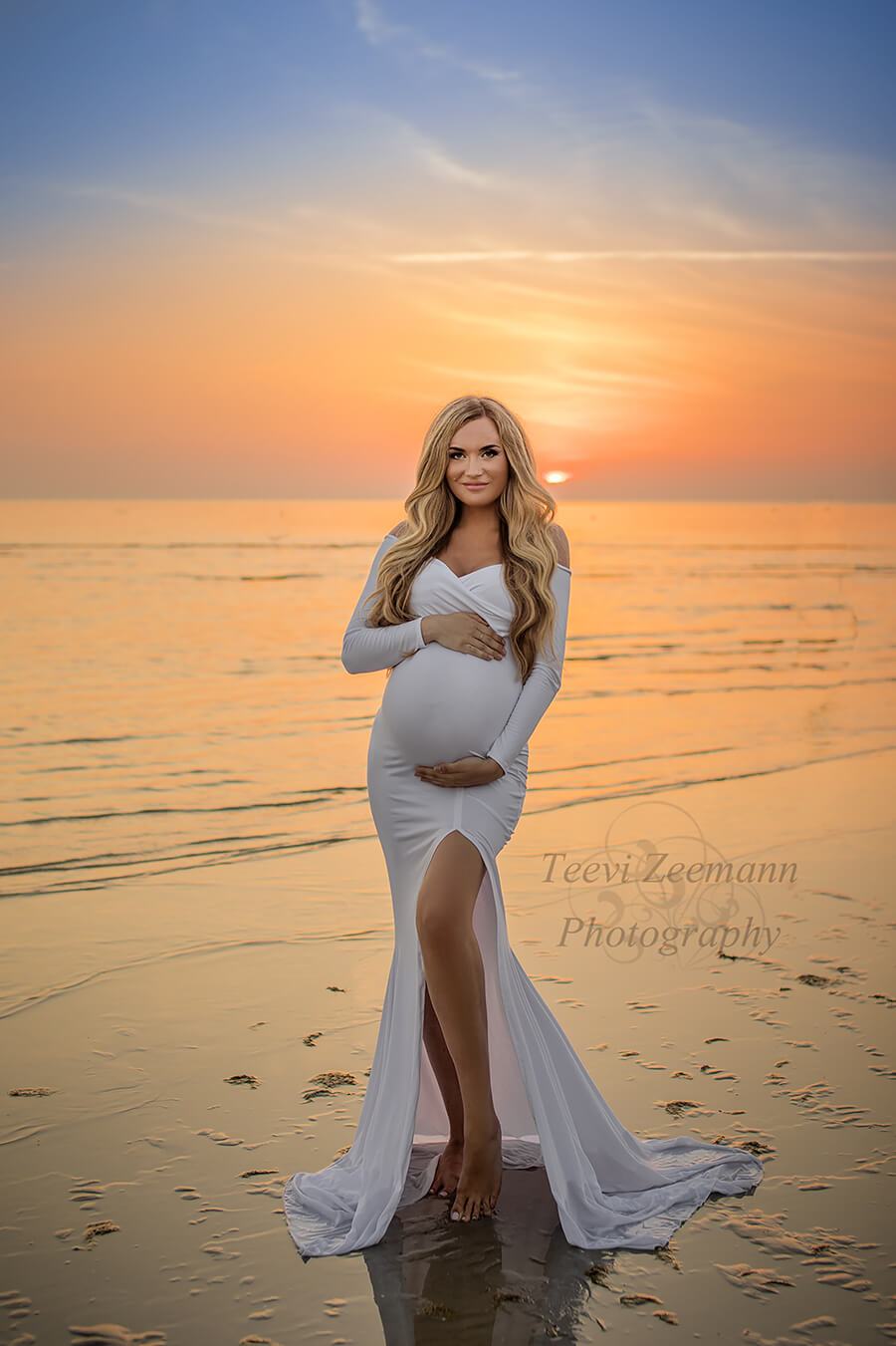 Blond pregnant model poses at the beach wearing a long tight white dress with a split on the skirt, long sleeves and sweetheart top. Model stares at the camera. She holds her belly with both hands.