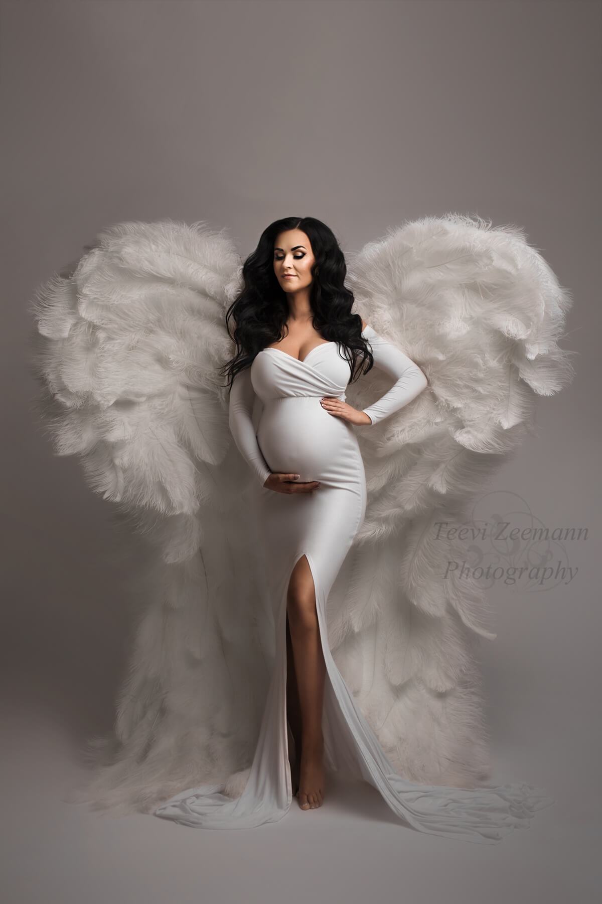 Brunette pregnant model poses in a studio wearing a long tight white dress with a split on the skirt, long sleeves and sweetheart top. Model has the eyes closed and holds her belly with one hand and her waist with the other. She has white wings on her back. 
