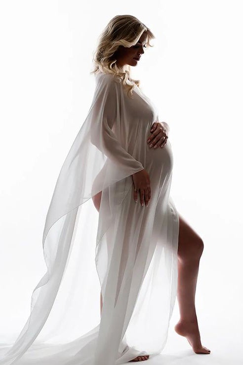 Blond pregnant model poses on her side wearing a white and transparent chiffon cape. She has her eyes closed and holds her belly. 