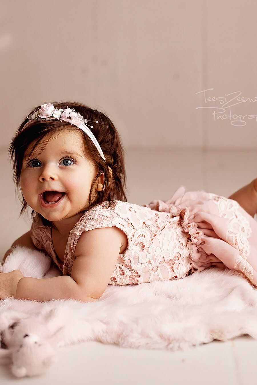 Little girl poses with a lace outfit in dusty pink color and a flower hair band. She is smiling and laying on a fluffy carpet. 