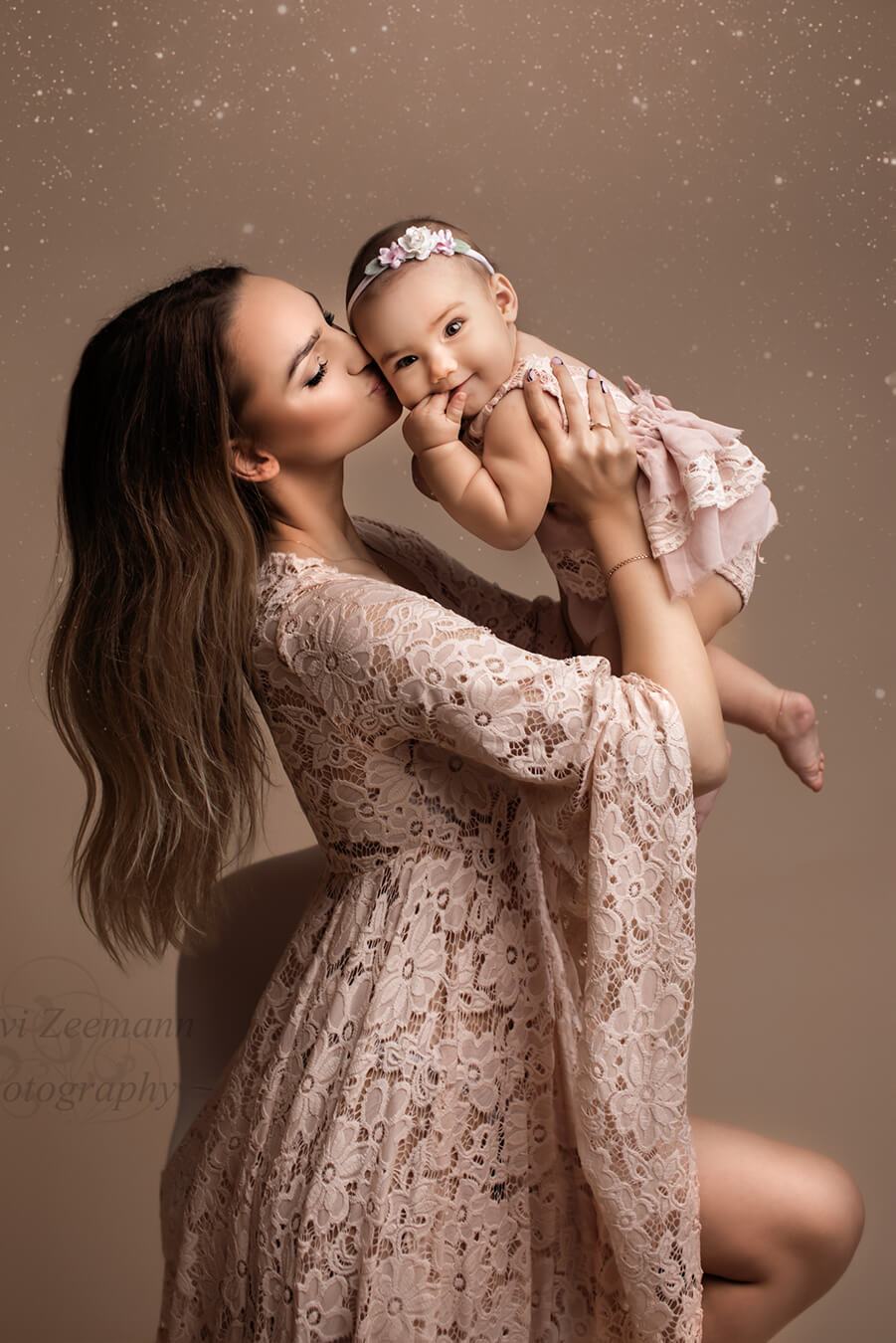 Photo session between mother and daughter. The mother has long brown hair and is holding her baby girl whilst giving her a kiss. The little girl stares and smiles at the camera. They are both wearing dresses in dusty punk color made of lace. The baby has a delicate head band with flowers. 