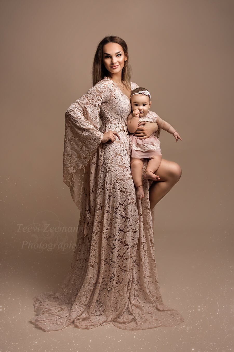 Photo session between mother and daughter. The mother has long brown hair and has her child sitting in one of her legs. They are both staring at the camera and wearing matching outfits in dusty pink. The mother wears a long dress with long kaftan sleeves made of lace and the baby a lace romper with a matching chiffon skirt - and a little head band with flowers. 