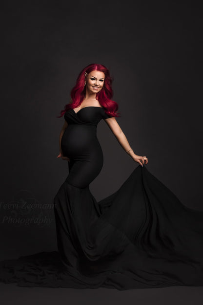 Pregnant model with bright, pink hair posing in a photography studio. She wears a black maternity dress with off shoulder sleeves and a mermaid skirt made of chiffon. The rest of the dress is made of jersey fabric which is tight fitting to her body. One hand is holding the skirt and the other one is under her belly. She is smiling and looking at the camera.