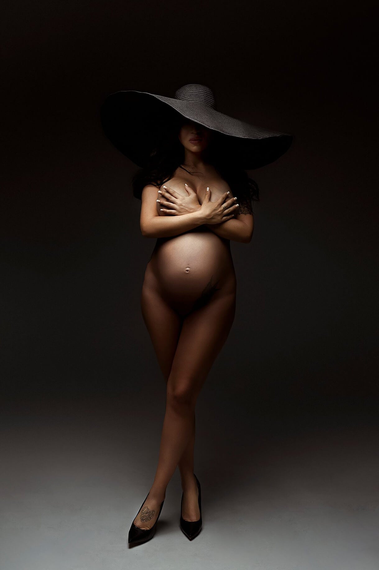 The woman is standing inside a studio. The product in the photo is a big black hat. She is wearing it on her head. The hat is covering up a part of her face. She is standing with her legs crossed and wearing high black heels. 