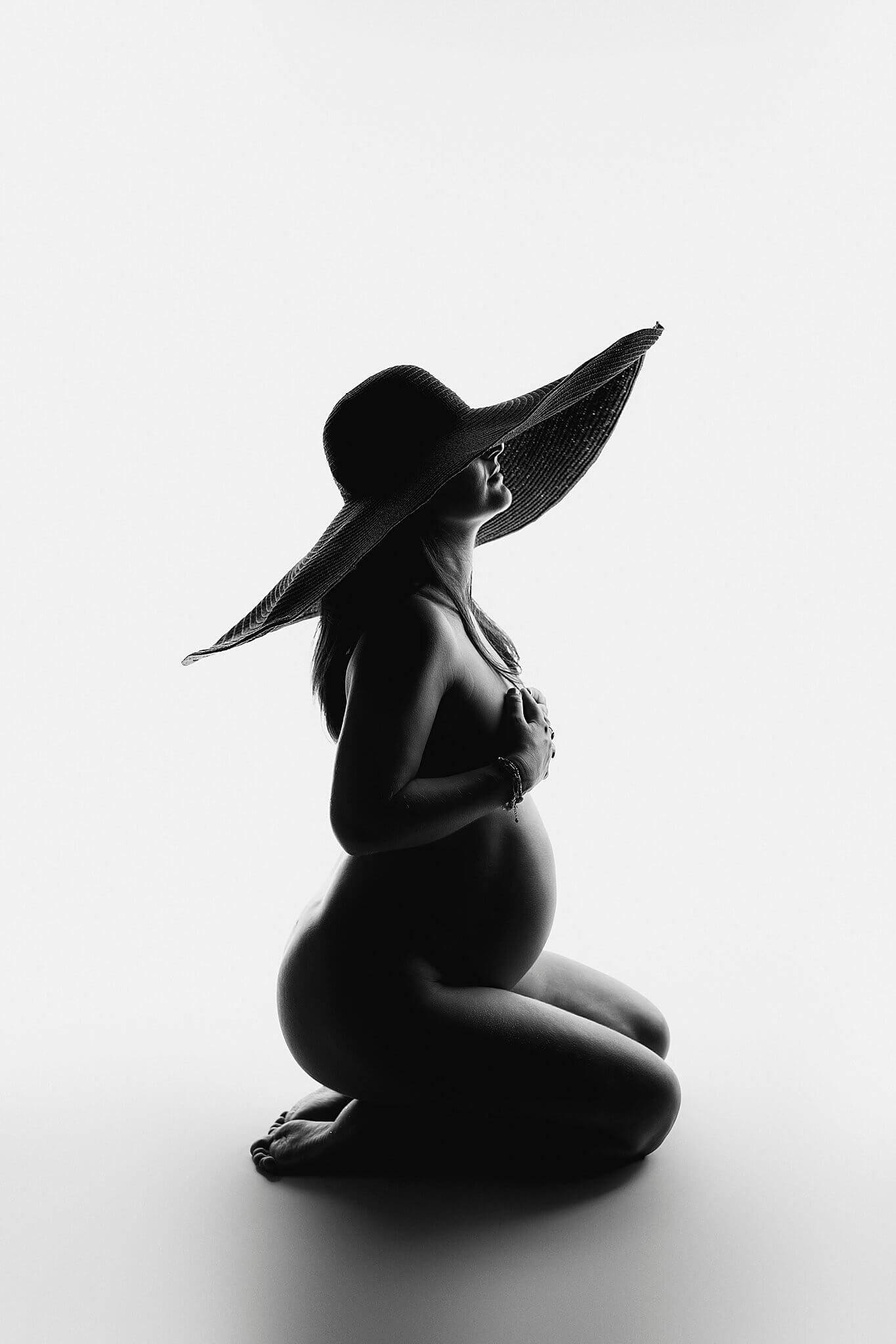 This is a black and white picture of a pregnant woman. The woman is sitting down on her knees and has a big black hat on her head. The hat is a real statement piece. 