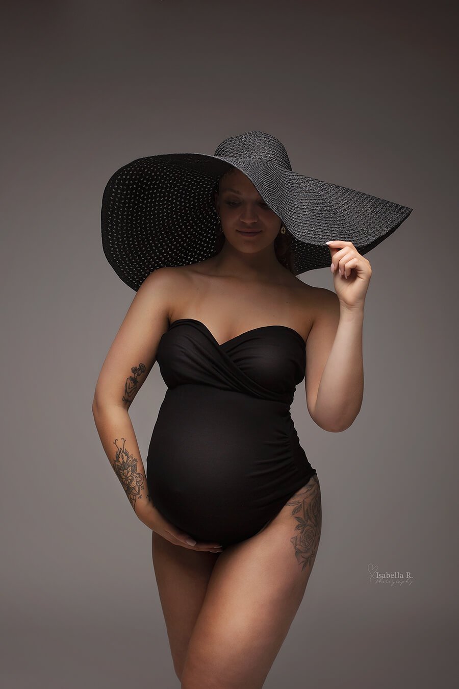 A pregnant model is wearing a black sleeveless bodysuit. The model has also a big black fashion hat on. The hat is perfect to use in your studio photography. She has a few tattoo&