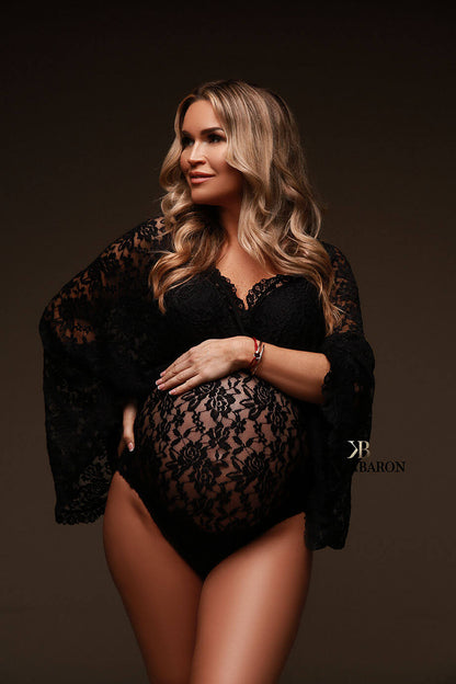 Blond pregnant model wears a maternity bodysuit during a photoshoot. The bodysuit is made of lace and features a low-v cut and kaftan sleeves top. The model looks to the side while holding her bump and waist. The photo is taken in a studio.