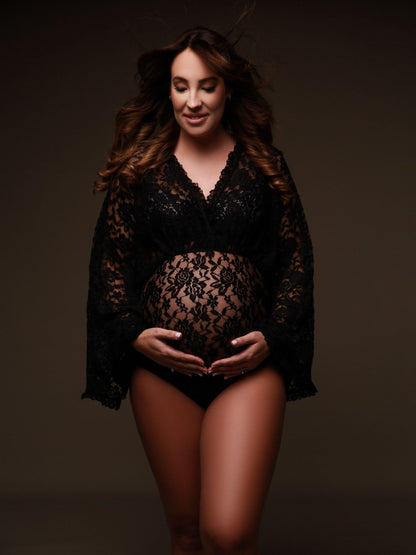 The model is pregnant and wearing a lace bodysuit. The top of the bodysuit is in a boho style. The woman has two hands underneath her belly. She has a soft smile on her face. The bodysuit is transparent. 