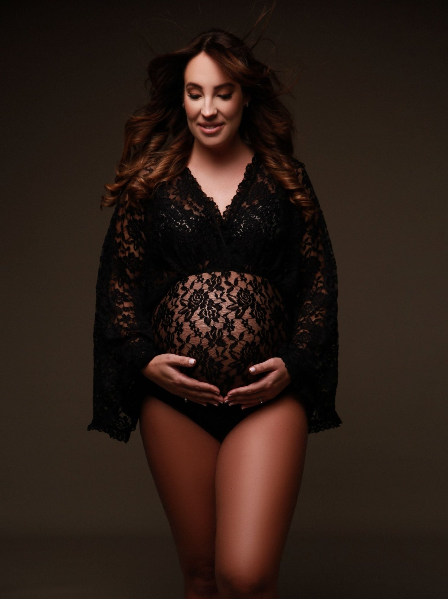 The model is pregnant and wearing a lace bodysuit. The top of the bodysuit is in a boho style. The woman has two hands underneath her belly. She has a soft smile on her face. The bodysuit is transparent. 