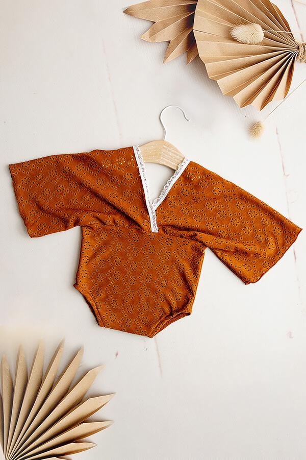 This is a product picture of our BOHO Brocant Cognac. The romper is made of Embrodery Lace and has a BOHO top with long wide sleeves. The neckline is finished with lace. In the corner of this picture you can see flowers for decoration.