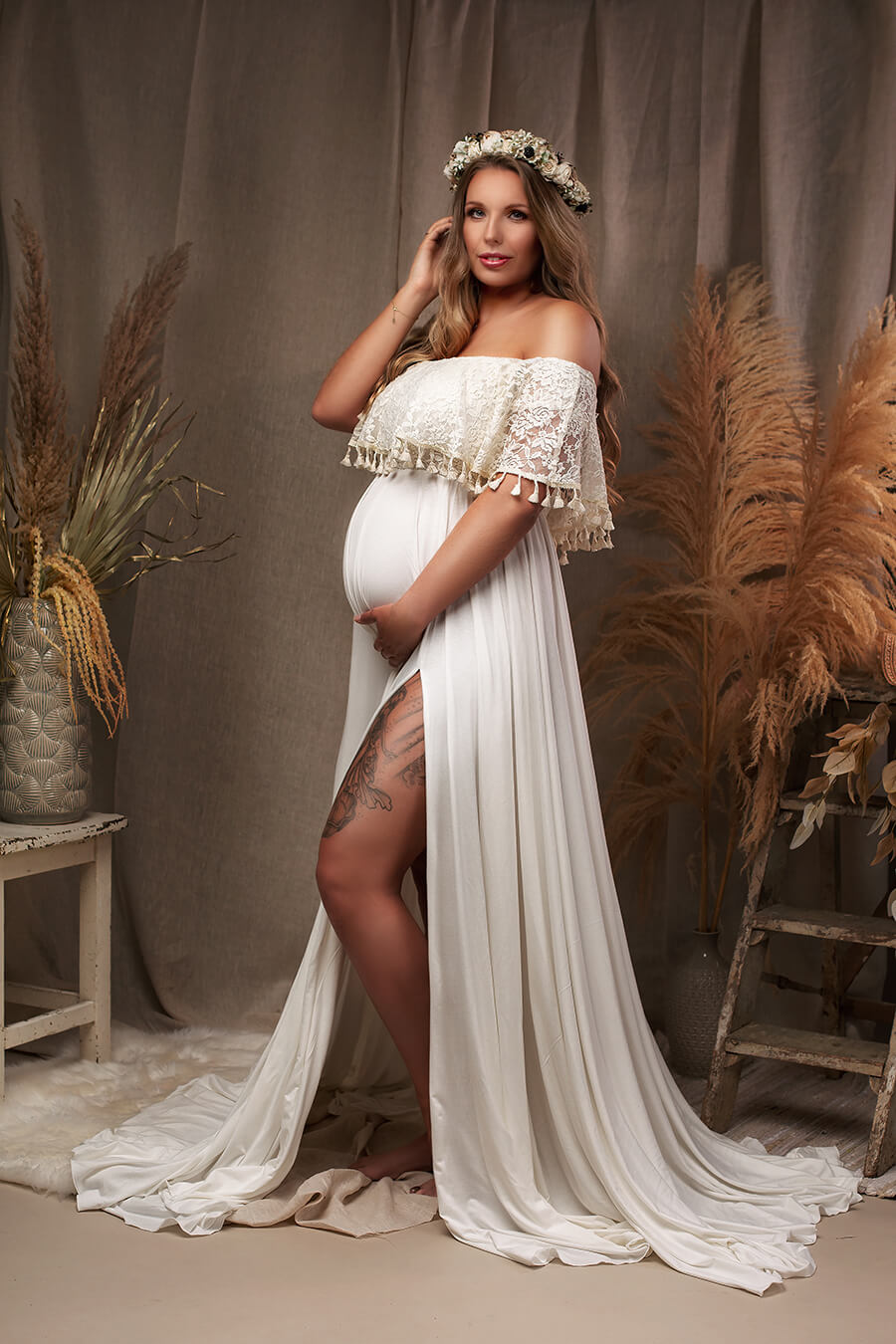 Blond pregnant model poses in a studio with the Boho Catania Maternity Dress with a flower gown. The photo shows the whole dress from a side view - a strapless top with a long circle skirt. The biggest detail on the picture is the side split from the dress, where you are able to see one of the legs from the model. The model faces the camera and holds her bump with one hand and her hair with the other. The top is made of bridal lace with brocante details at the ending.