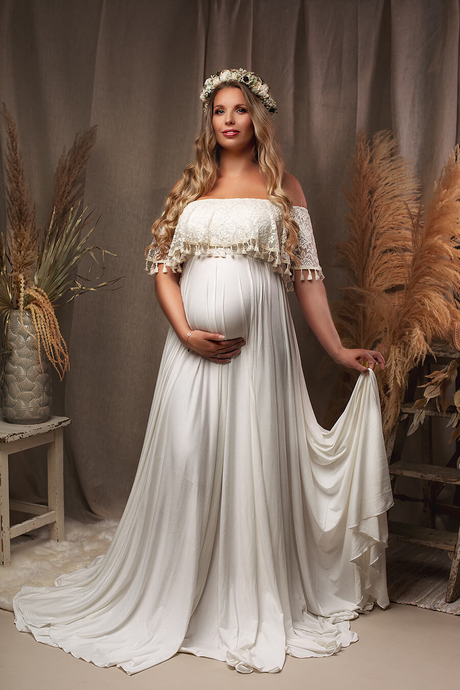 Blond pregnant model poses in a studio with the Boho Catania Maternity Dress with a flower gown. The photo shows the whole dress - a strapless top with a long circle skirt. The model faces the camera and holds her bump with one hand and the skirt with the other. The top is made of bridal lace with brocante details at the ending.