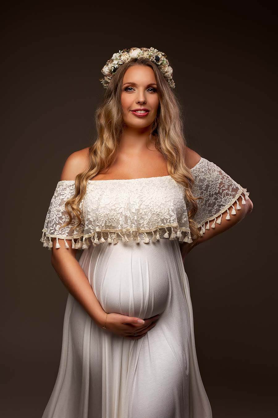 Blond pregnant model poses in a studio with the Boho Catania Maternity Dress with a flower gown. The photo is a close up on the top from the dress and a split on the side. The top is strapless with elegant lace and vintage brocante details at the ending.