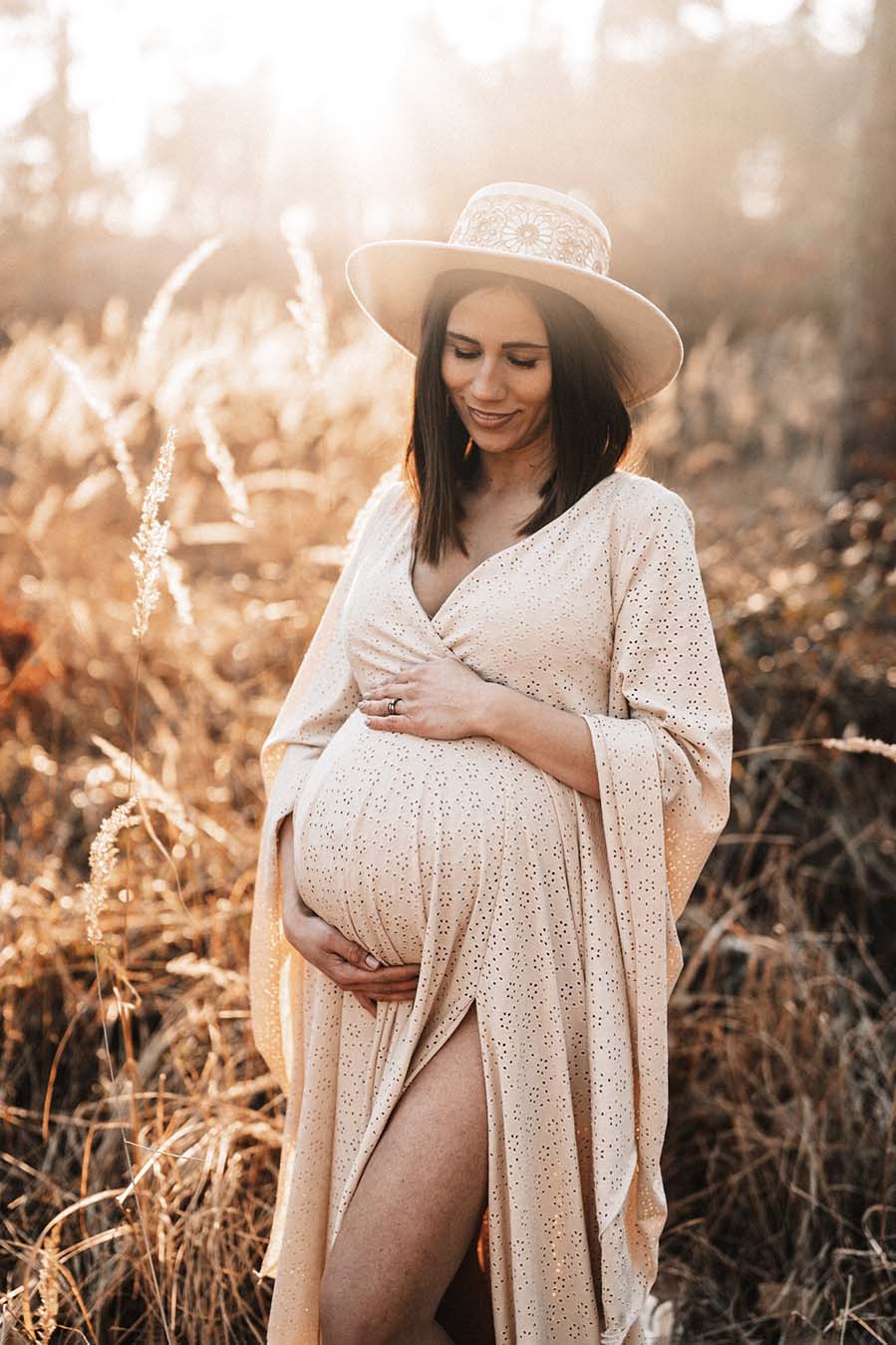 model poses outside by a field wearing a long dress made of jersey with a low v cut neckline and kaftan sleeves. the dress is made in sand color. she has a boho styled hat to match the look.
