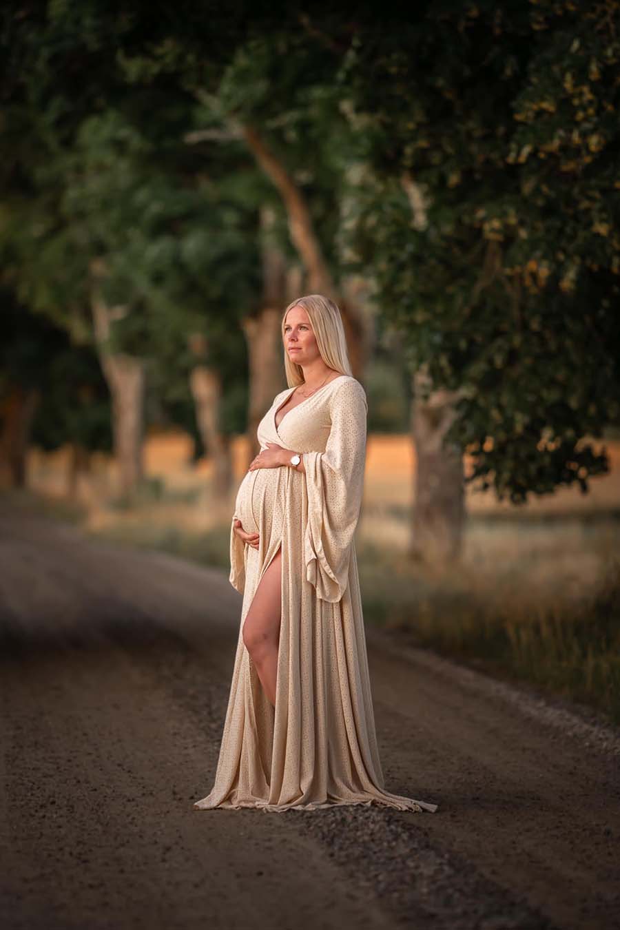 blond pregnant model poses on a road wearing a long sand dress made of jersey. the dress features a low v cut neckline and a circle skirt with a delicate side split/ she poses on her side, with open eyes and holding her baby bump with both hands.