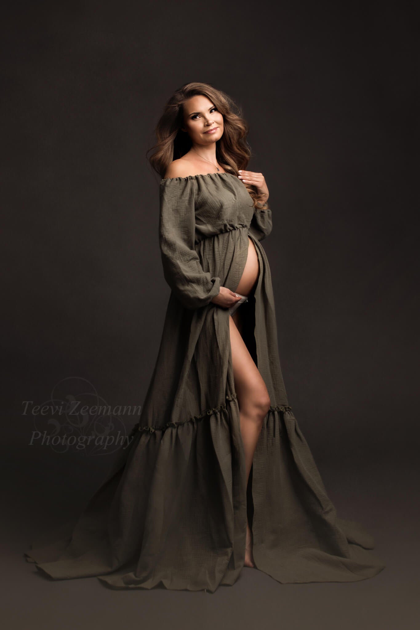 Dark blond pregnant model poses in a studio wearing a mii-estilo boho style dress in olive color. She stands facing the camera but her body is turned a bit to the side. Her bump and one of her legs can be seen through the split on the skirt. She holds her hair with one hand and her bump with the other one. 