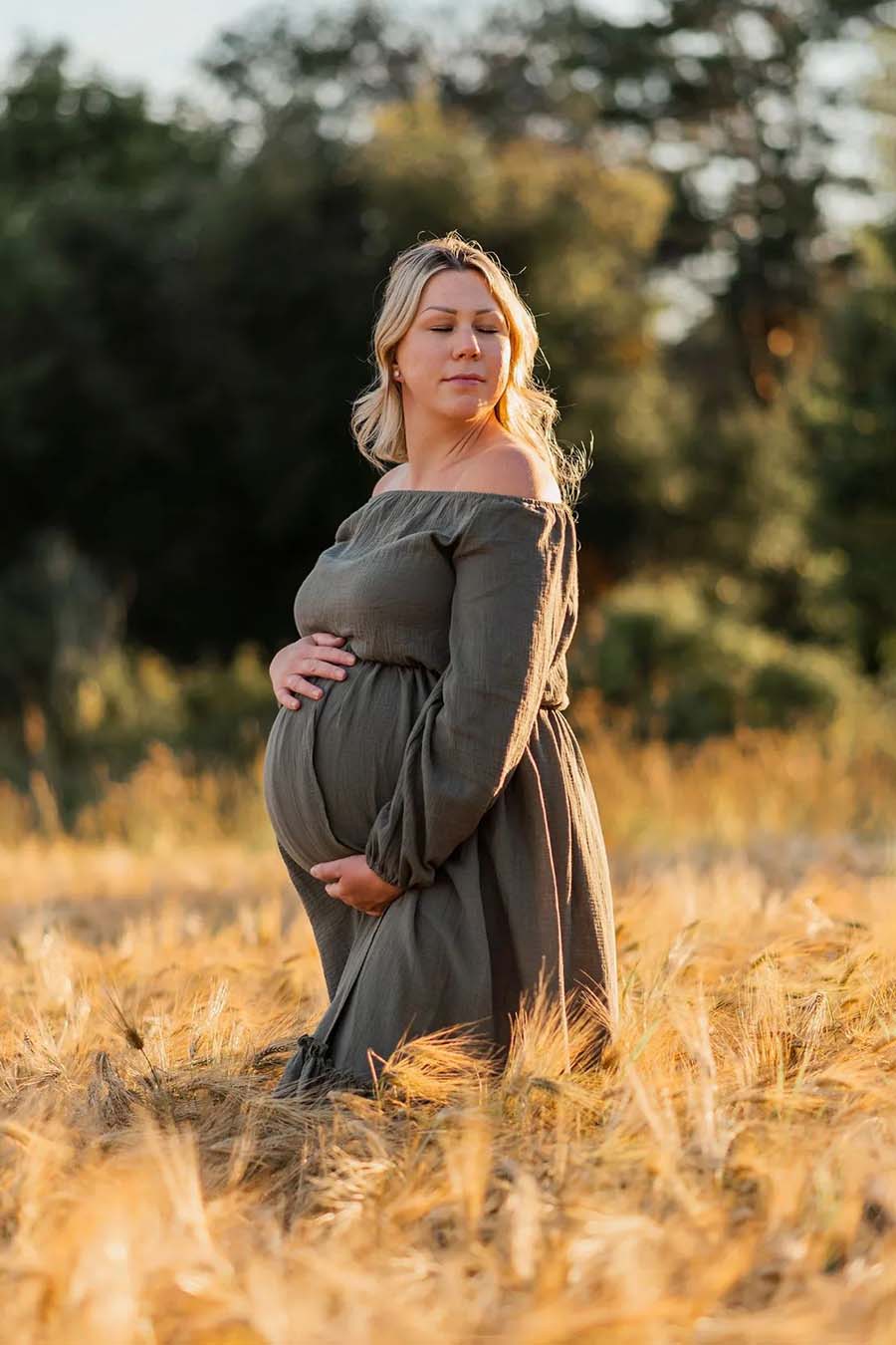 blond pregnant woman poses outside with her eyes closed wearing an olive dress with off shoulder neckline. 