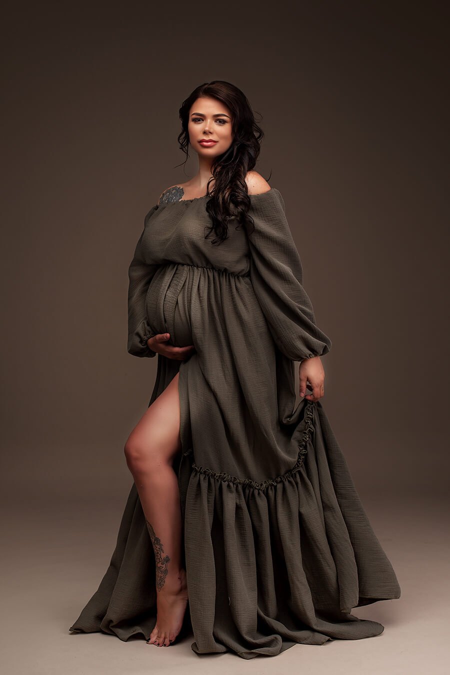 Brunette model poses for a maternity photography session with a pregnancy boho style Mii-Estilo dress named Ibiza. The dress has long puffy sleeves and ruffle details in the long skirt. The skirt has also an opening that can be seen in the photo. The model poses showing one of the legs through the split and her hands are holding her bump and the skirt. Model faces the camera and the whole dress can be seen in the photography. 