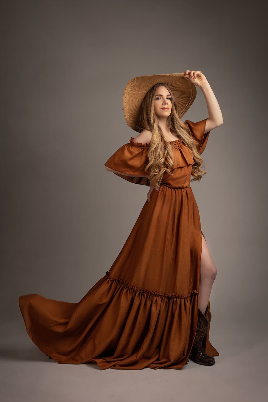 blond model pose sin a studio during a boho chic photoshoot. she has a long cognac dress with ruffle details and a brown hat to match the style. 