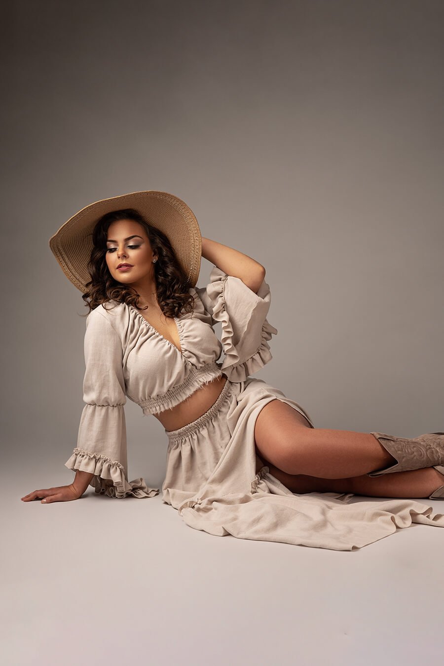 brunette model poses in a studio for a boho western chic photoshoot. she wears a ecru color set with a lot of ruffles on the top and the skirt. she wears boots to match the style. the model is sitting on the floor and leans the body on 1 arm. the other holds her hear, where she has a brown hat to match the style.