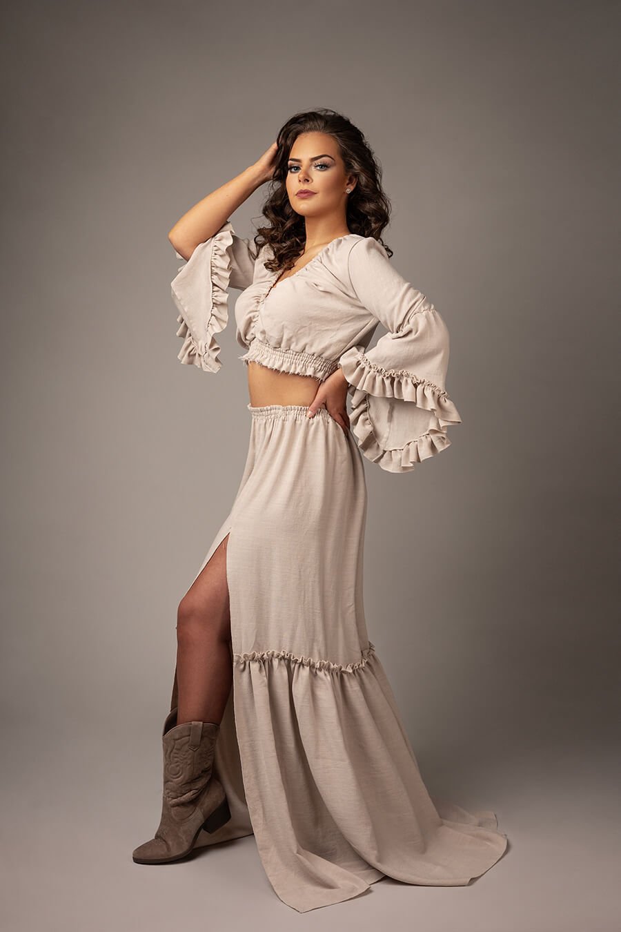 brunette model poses in a studio for a boho western chic photoshoot. she wears a ecru color set with a lot of ruffles on the top and the skirt. she wears boots to match the style.