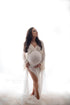 Brunette pregnant model poses in a studio during a maternity photoshoot. She has her eyes closed and her head facing up. She wears an off white long dress made of lace and chiffon and stands in tulle fabric. One hand holds the skirt and the other holds her bump,