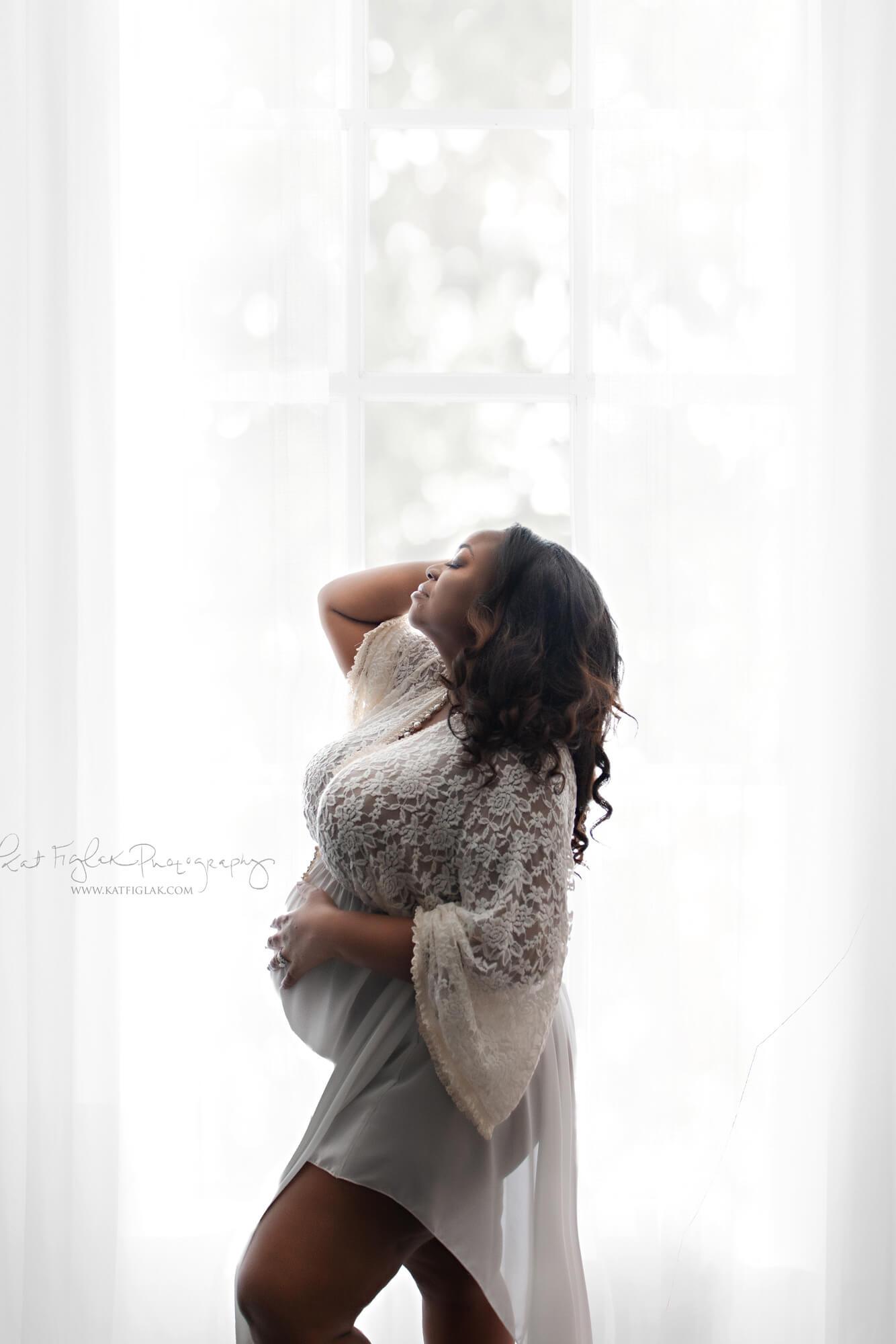 Pregnant model poses in studio during a maternity photosession. She holds her head with one hand and her belly with the other. She wears an off white long dress made of lace and chiffon. Her eyes are closed and facing up. 