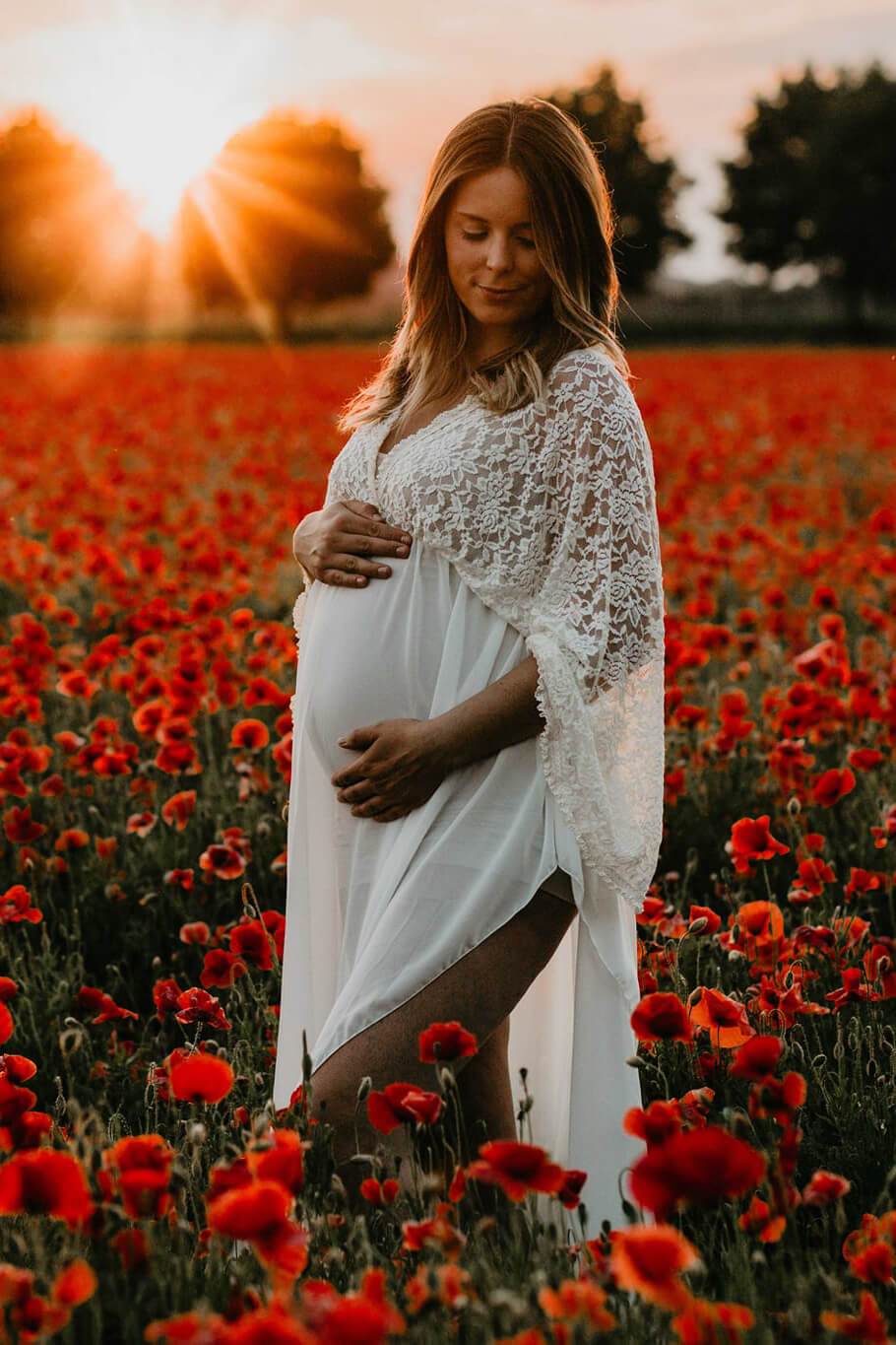 Blond pregnant model poses outside in a flower field wearing an off white long dress made of lace and chiffon. She has her eyes partially closed and looks down while holding her bump with both hands. 