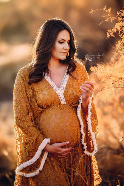 Pregnant model poses outside with a cognac dress made of lace with details in white color on the sleeves and top. The top has a low v cut neckline and kaftan sleeves. She is looking to the side and holds her bump with one hand and some flowers with the other one.